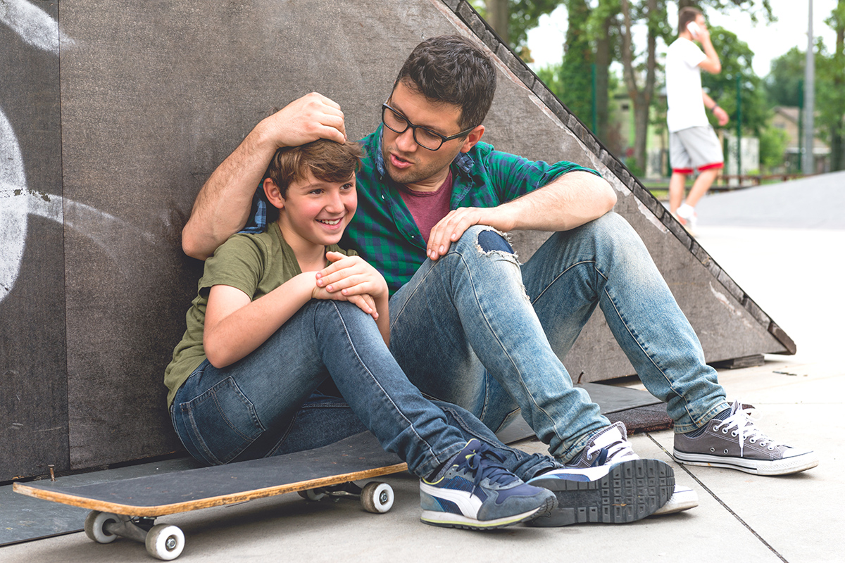 A dad sitting outside with is son who is sitting on a stakeboard.