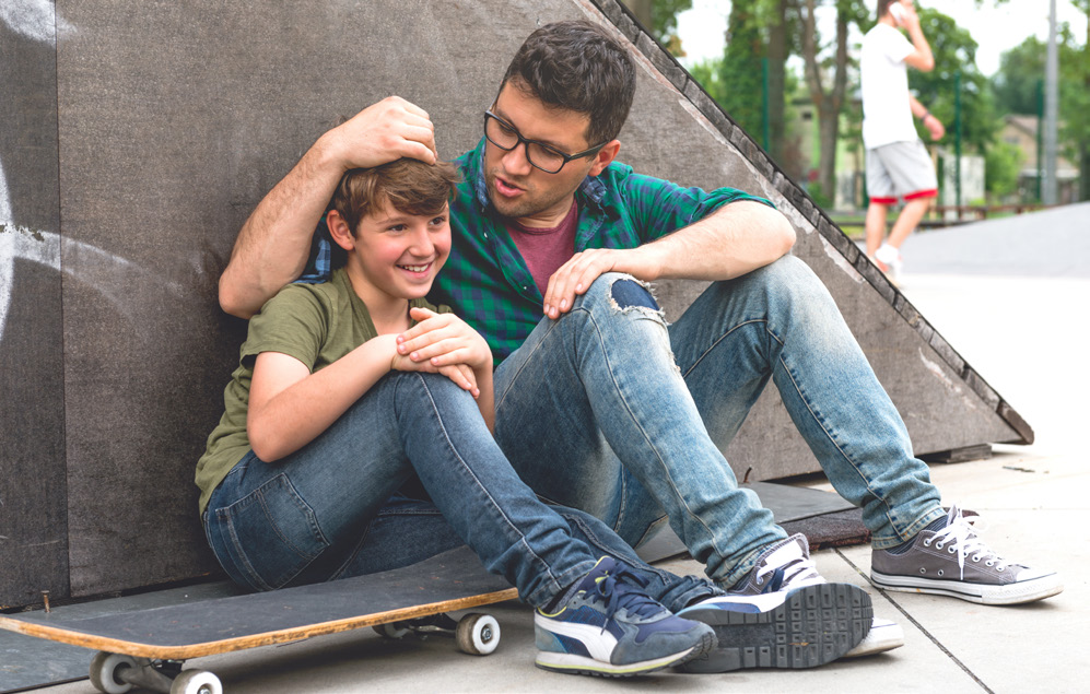 A father is sitting down with his kid and his skateboard.