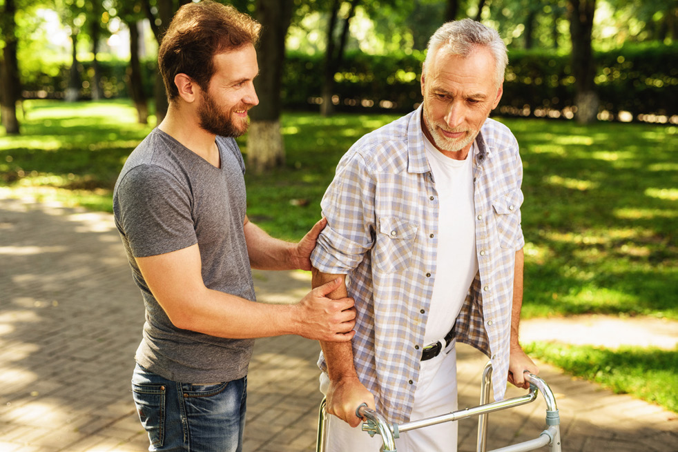 A younger gentlemen helps an older man using a walker. 