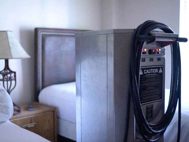 A heat treatment machine sits inside a bedroom with wooden furniture and white sheets. The machine is silver and rectangular with a black, flexible hose. One side of the machine has a black sticker saying, "Caution" below several buttons that are lit up red and yellow.