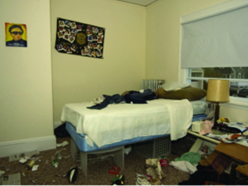 A bedroom with a bed with white sheets and clothing items thrown atop it sits next to a small nightstand with a cream colored lamp and several items of trash and clothing. Under the bed are items stored and jumbled up. The floor is carpet and cluttered with paper trash, clothing, and other random items. The walls are a cream color with two posters hung, one showing a face, the other random shapes, which is crooked both are very colorful.