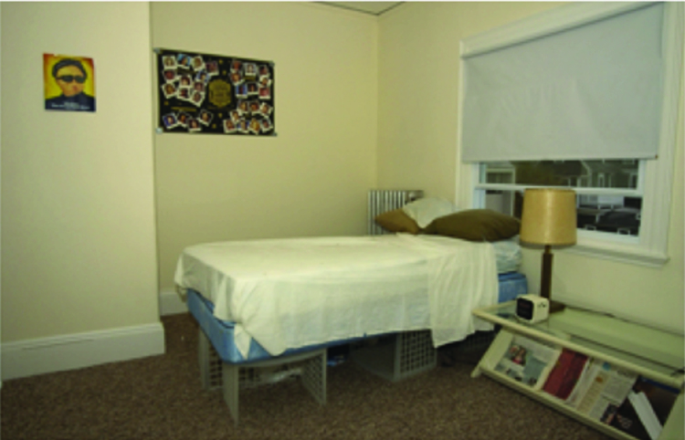 A bedroom with a neatly made bed with white sheets sits next to a small nightstand with a cream colored lamp. Under the bed are items stored and organized neatly. The floor is carpet and clean with no clutter. The walls are a cream color as well with two posters hung, one showing a face, the other random shapes both are very colorful.