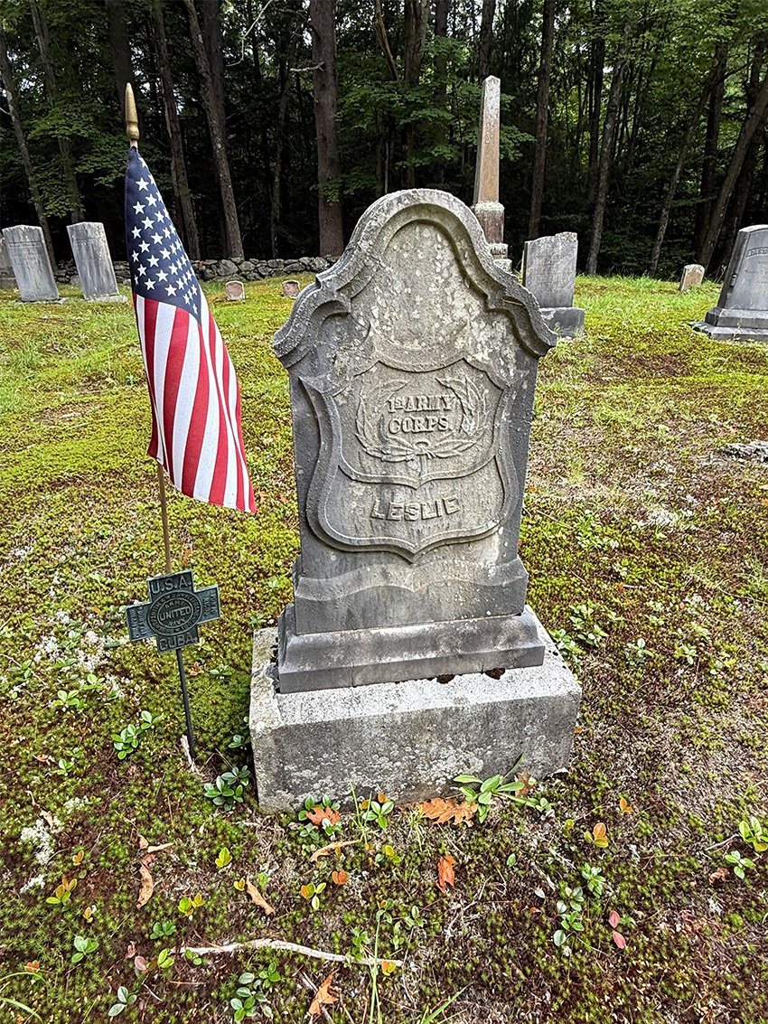 Weathered gravestone with American flag in quiet wooded cemetery, mossy ground and surrounding historic headstones visible.