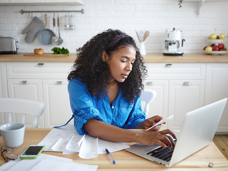 A lady sits at a wooden table typing on a laptop. She holds a piece of paper and there are various other papers atop the table next to the laptop. There is also a cell phone and a white coffee cup next to the paper pile.