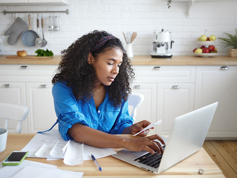A young woman doing bills on a lab top on the kitchen table.