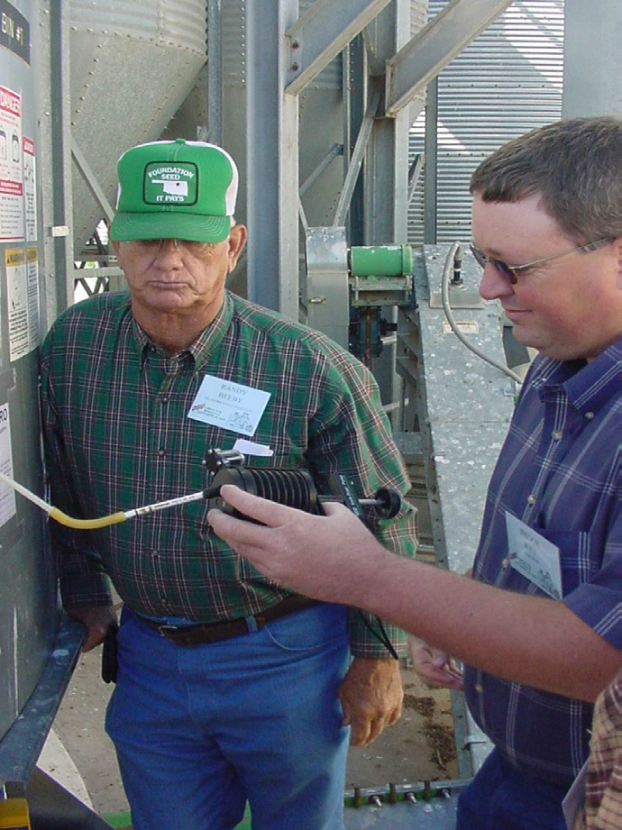 Two people standing by a grain bin with a device attached to the bin. Two people standing by a grain bin with a device attached to the bin.
