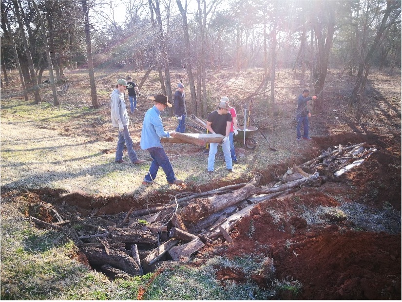 A group of 7 people near a hügelkultur, filling a curved trench with logs and wood debris as the base of a hügelkultur bed in red clay soil.