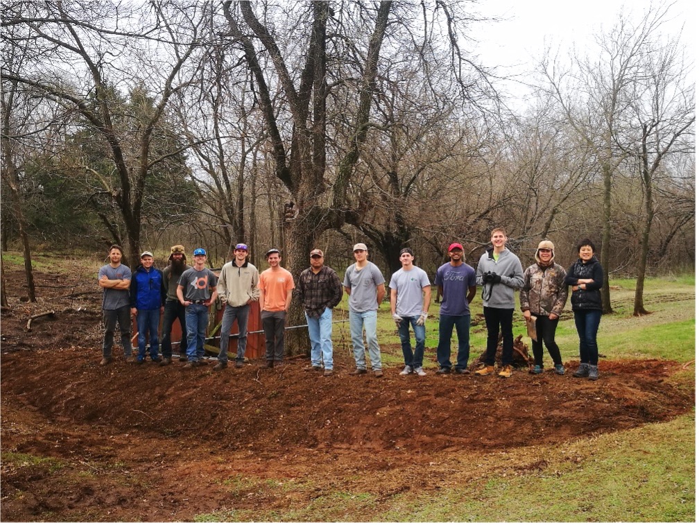 Figure 14 Sustainable Landscapes HLA-6478 13 people standing on a large soil mound in a wooded area wearing casual outdoor clothing with bare winter trees in the background.