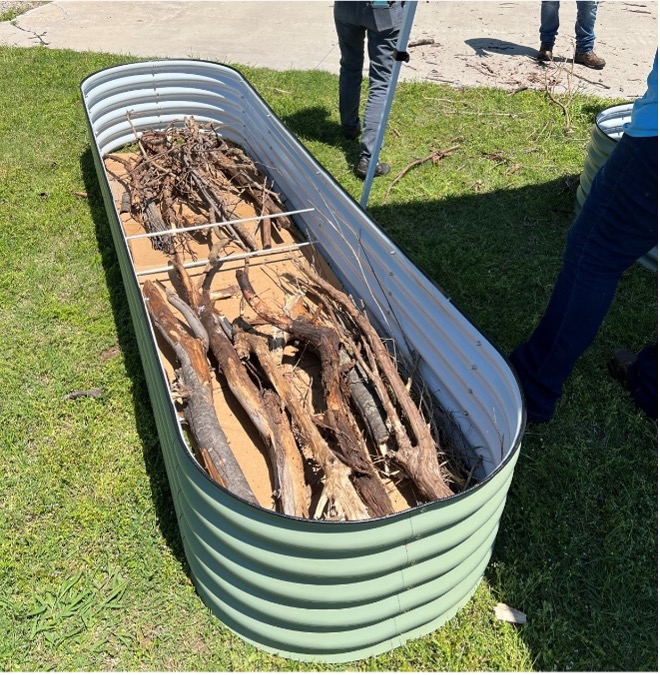 Oblong metal raised garden bed partially filled with cardboard logs and branches showing early hügelkultur variation on grass.