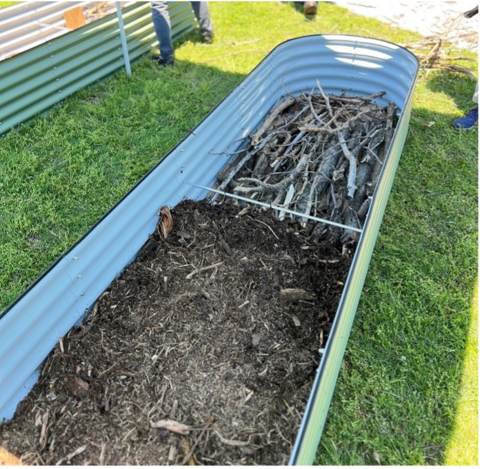 Oblong metal raised garden bed showing layered branches on one end and mulch/compost on the other for hügelkultur variation.