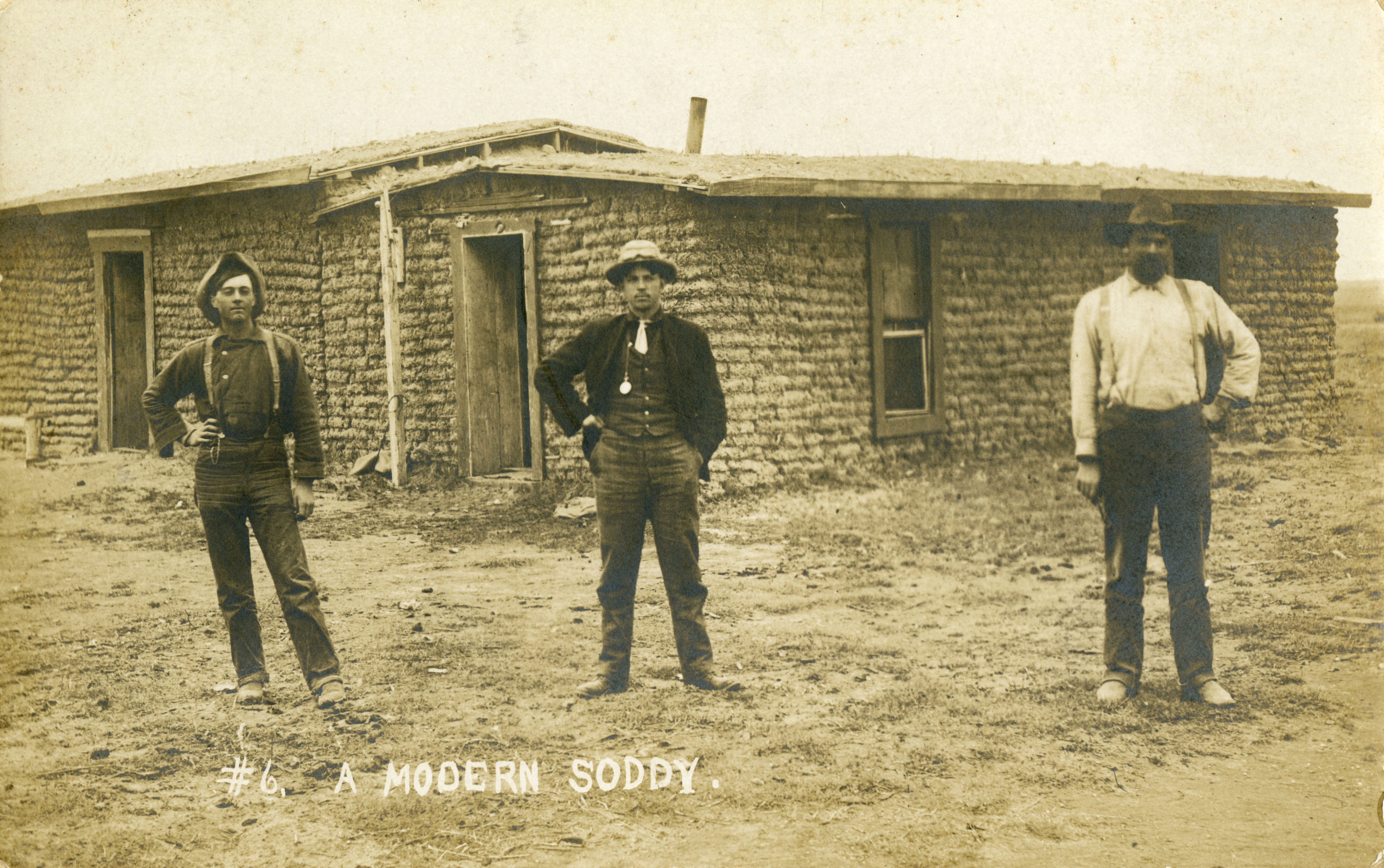 A postcard of three men standing in front of a sod house.