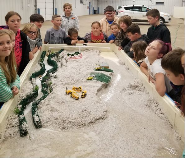 A sandbox with toy vehicles and stream trailer as children observe around the table of sand.