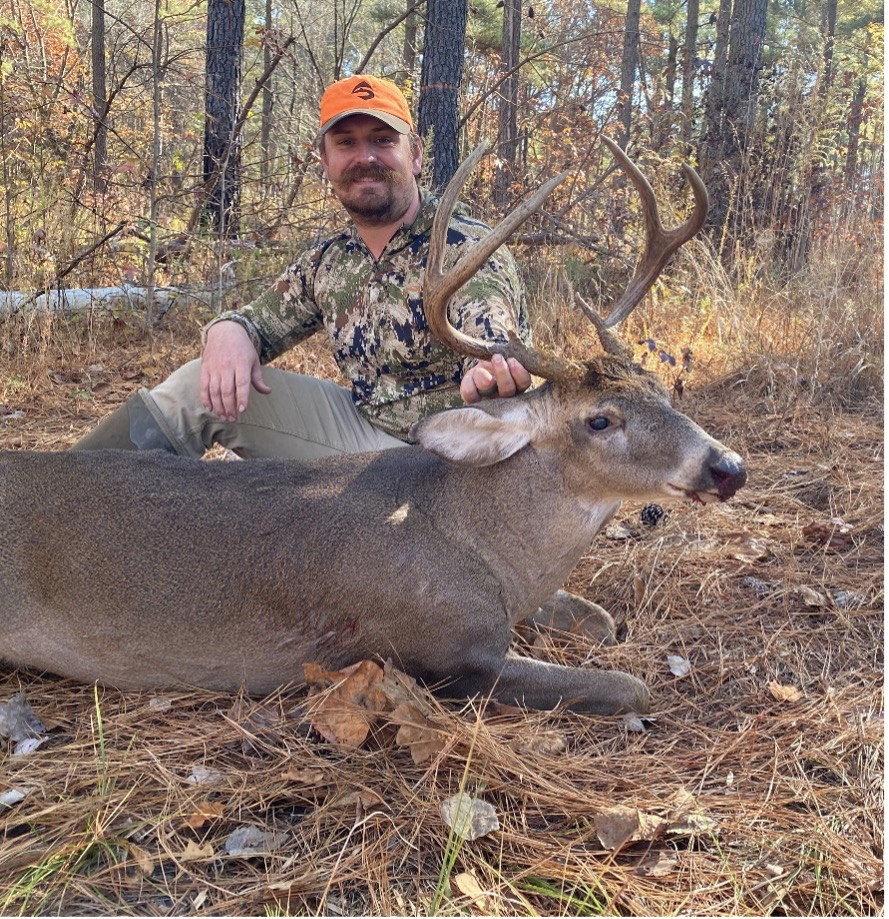 A male in an orange hat and camo holding a buck by the antlers in the middle of a bare forest.