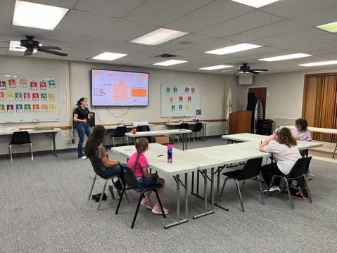 A presenter in the front of a classroom of children sitting at a table.