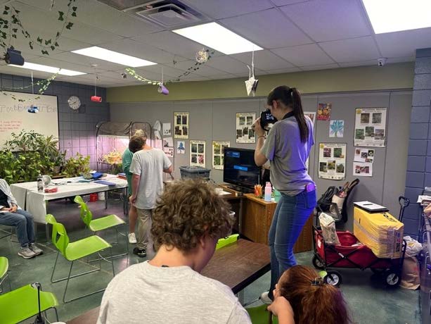 A girl is standing on a chair in the classroom using a camera to take a photo.