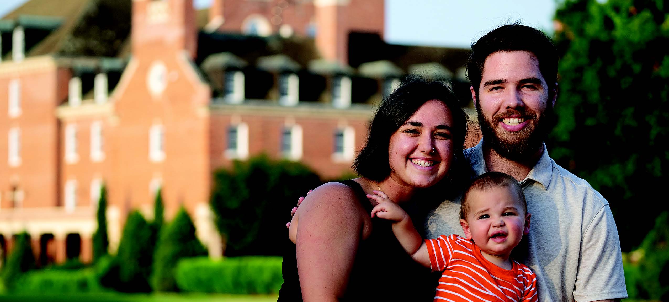 A man, woman and baby standing together, smiling. A building is in the backgroud.