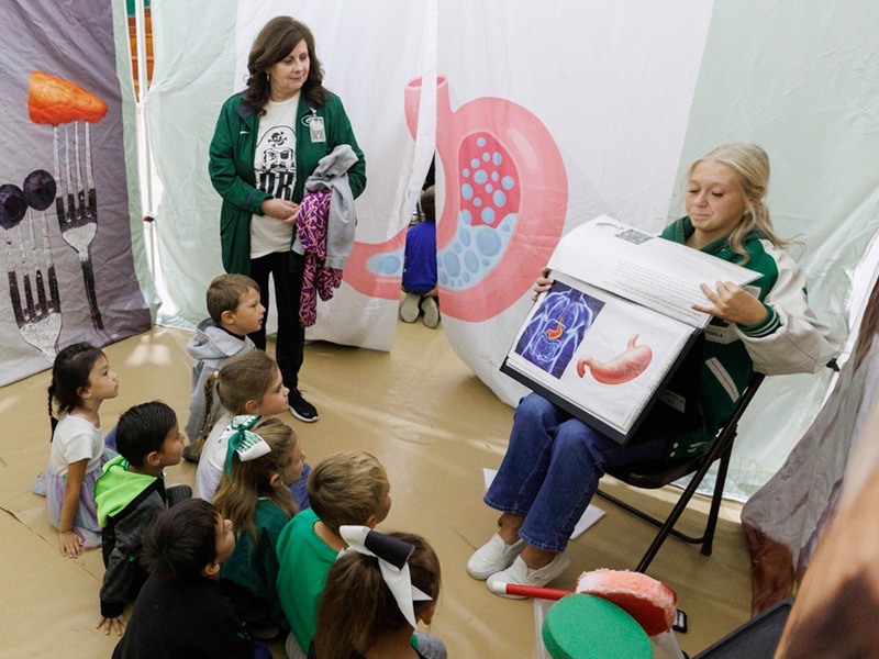 Young students sitting on the floor learn about the stomach from a Farm to You presenter.