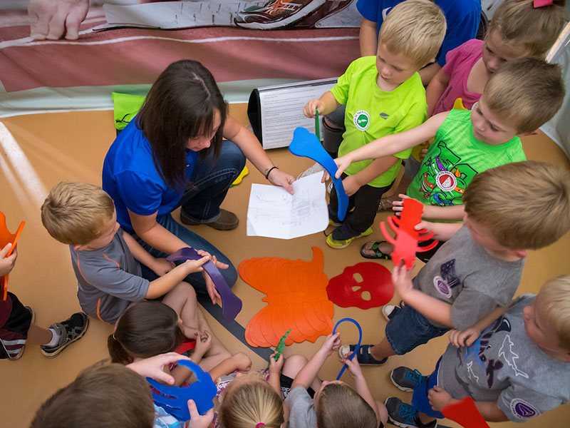 A female instructor kneeing on the floor with kids building a body out if stencils.