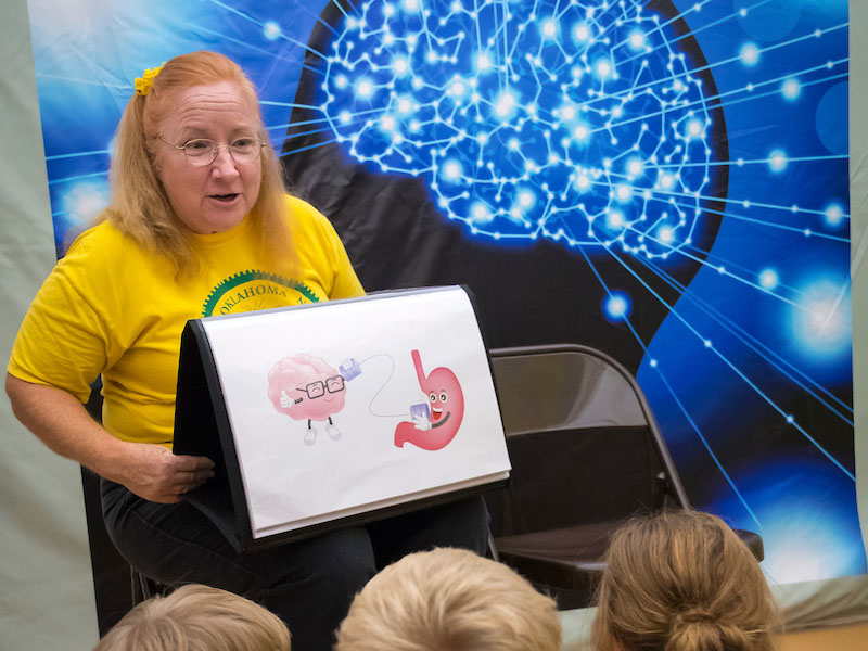 A female instructor sitting in a chair holding a flip out picture book in front of kids.