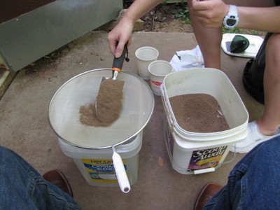 A spade moving sand from a bucket to be sifted through a screen sitting over an empty bucket. A spade moving sand from a bucket to be sifted through a screen sitting over an empty bucket.