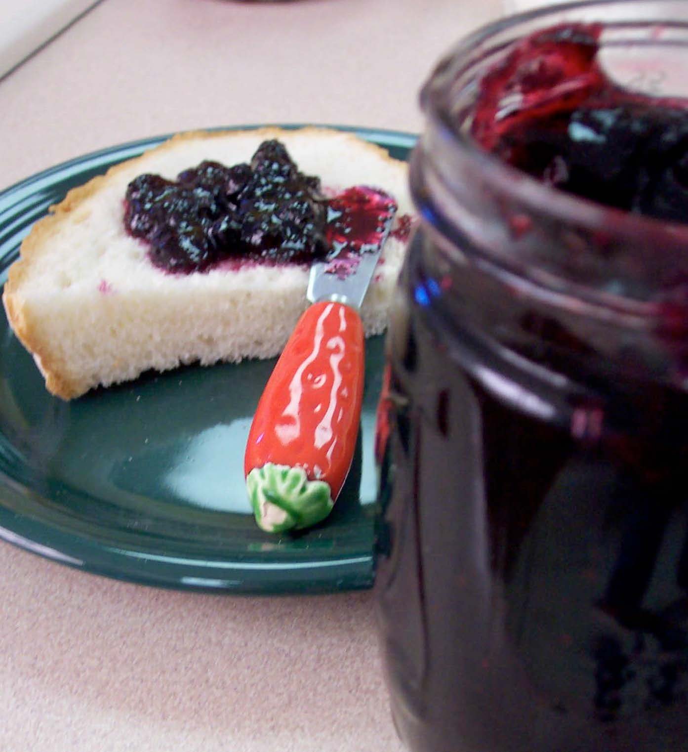 Blueberry jam spread on a piece of bread on a green plate on a table with the jam jar next to it.