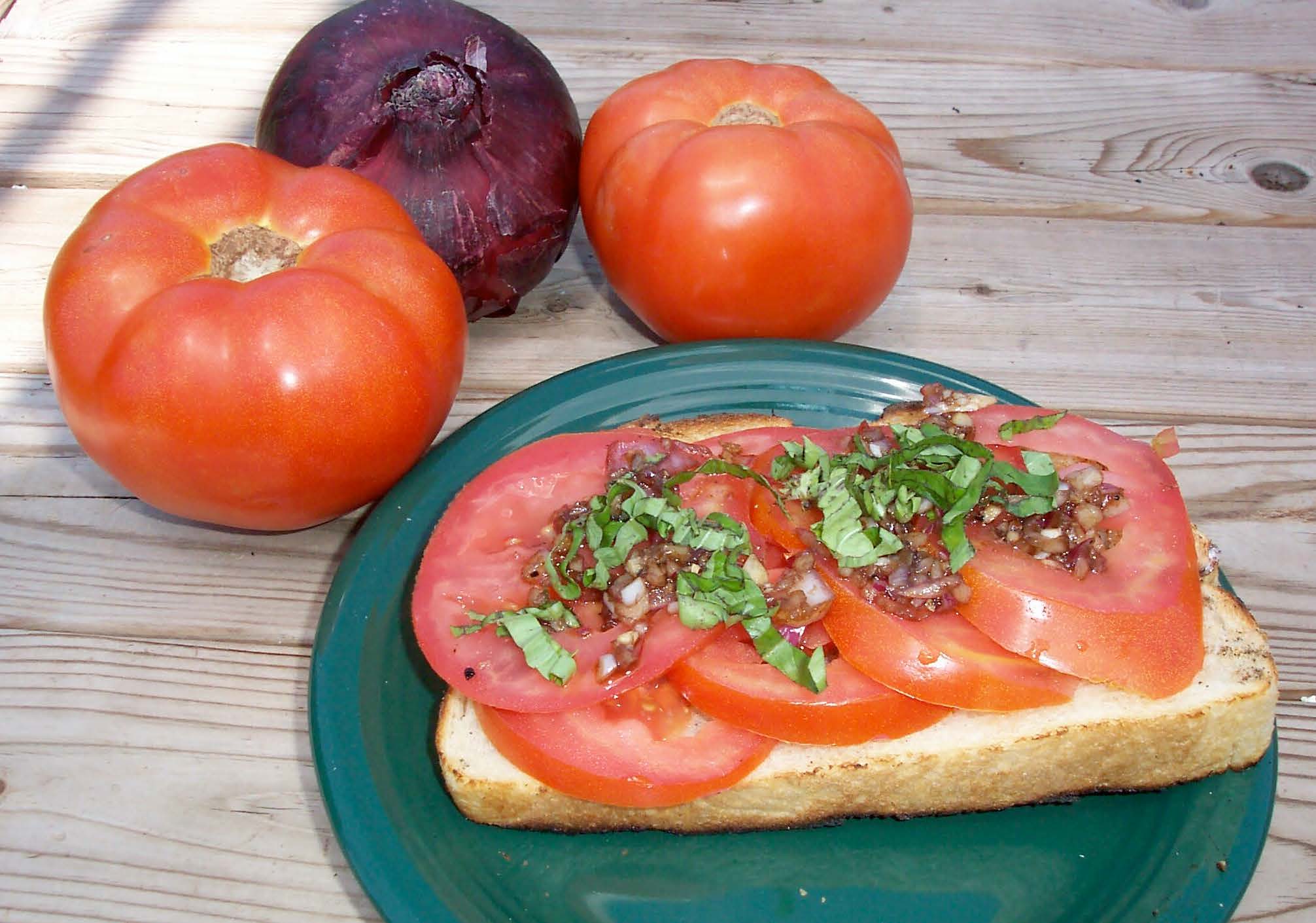 Grilled bread and tomatoes on a green plate on a wooden table with tomatoes and an onion in the background.