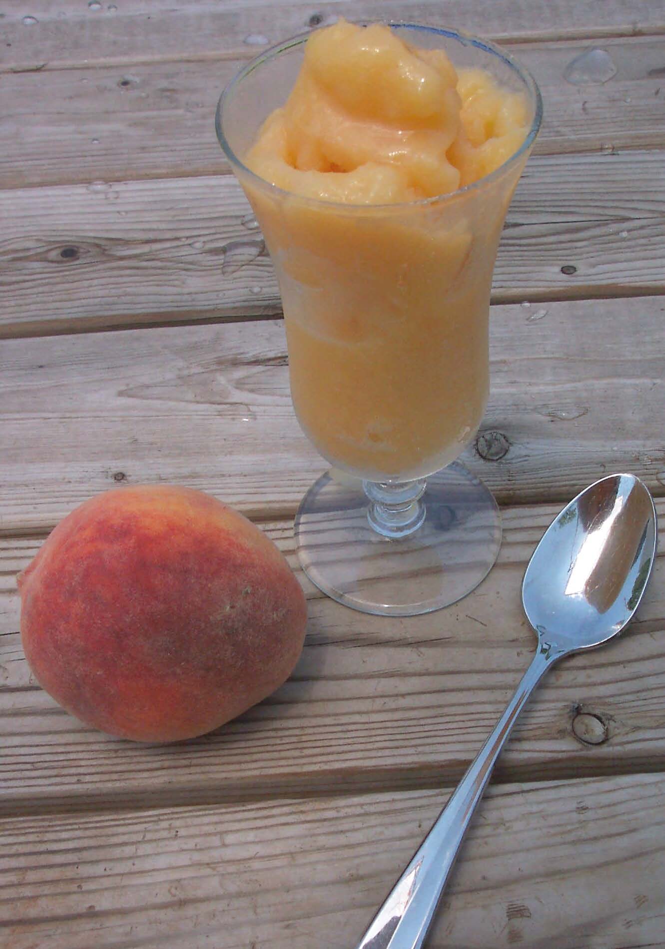 A clear glass full of orange sorbet on a wooden table with a peach next to it.