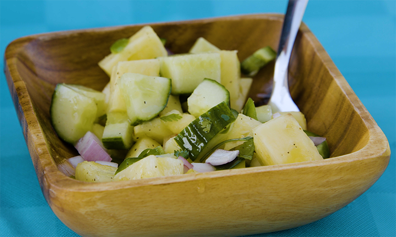 In a wooden bowl are cubed green cucumbers and yellow chunks of pineapple, covered in a clear sauce. A silver spoon rests on the right side of the bowl, partially under the salad. 