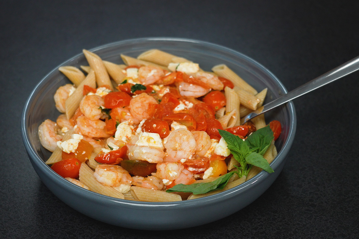 A bowl of pasta noodles, shrimp, and tomoatoes topped with feta cheese and basil leaves.