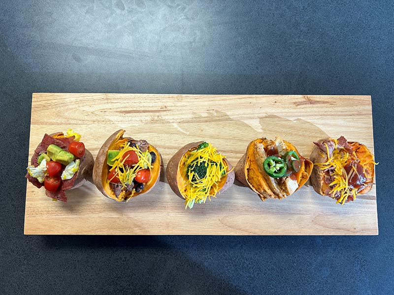 Open sweet potatoes in a row on a plate topped with various toppings like tomatoes, avocado, cheese, jalapeno and bbq sauce.