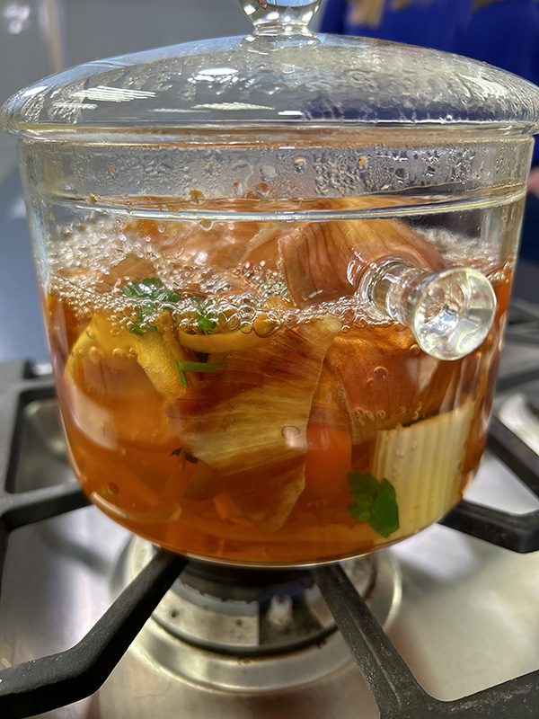 A pot of brown vegetable broth with vegetables in a clear pot on a stove burner.