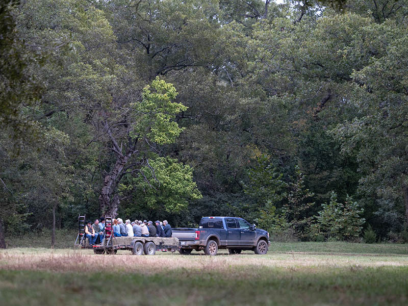 A truck pulling a flatbed trailer with people sitting on the trailer driving attendees to the field location for Native Pecan Harvest Field Day at Leon Bailey's Pecan Farm.